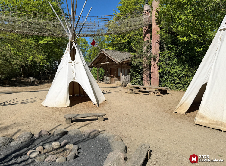 Tipi-Lager im Abenteuerland des Hansa-Park mit weißen Tipis, Feuerstelle, Holzbank und rustikaler Blockhütte im Hintergrund
