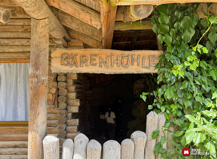 Eingang zur Bärenhöhle im Hansa-Park mit Holzschild und rustikaler Blockhüttenfassade, umgeben von grünen Efeuranken, ein Mädchen betritt den dunklen Höhleneingang