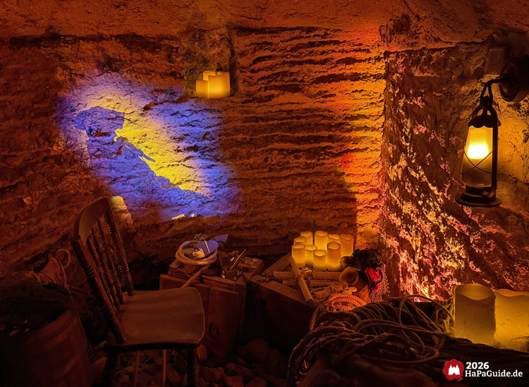Raum in der Bärenhöhle des Hansa-Parks mit Kerzen, Laternen und alten Gegenständen, beleuchtet durch gelbe und blaue Projektion, erzeugt die Atmosphäre eines Roanoke Seefahrers