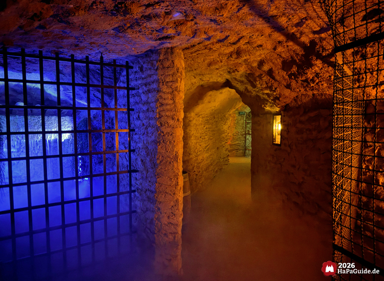 Unterirdischer Gang in der Bärenhöhle des Hansa-Parks mit bläulich beleuchtetem Gittertor und Nebeleffekt, daneben Laternenlicht, raue Steinwände und ein Wasserfall