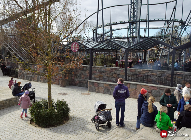 Besuchende schlendern an einem kühlen Tag über den Vorplatz der Achterbahn Royal Scotsman im Hansa-Park, vor der schwarzen Schienenkonstruktion Nessies mit Bahnhof hinter einer Steinmauer