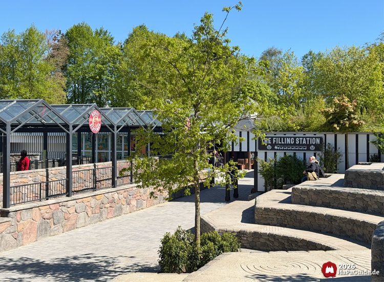 Sonniger Frühlingstag im Hansa-Park mit Blick auf den Bahnhof des Royal Scotsman und den Kiosk The Filling Station, umgeben von grünen Bäumen und Steinmauern