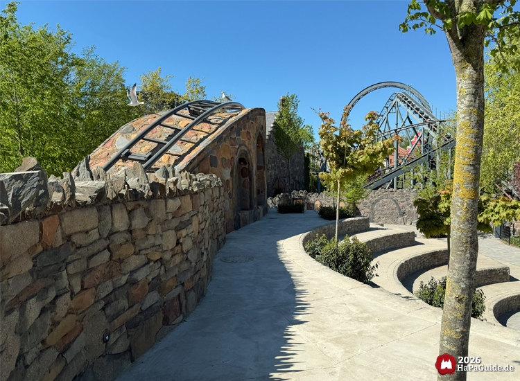 Steinterrasse mit geschwungener Mauer und Viadukt im Bereich Bezauberndes Britannien im Hansa-Park, umgeben von grünen Bäumen und dem markanten Looping Nessies bei sonnigem Wetter