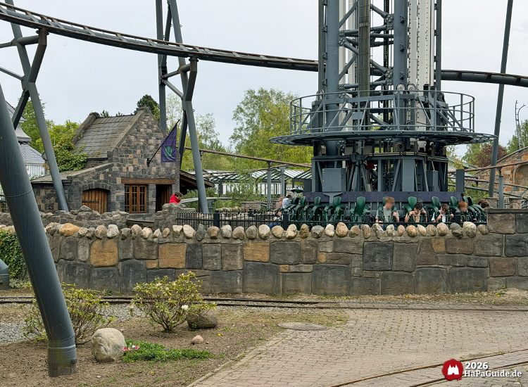 Blick auf den Highlander Gyro-Drop-Tower und die angrenzende Station der Achterbahn Royal Scotsman im Hansa-Park, umgeben von Steinmauern und grüner Vegetation an einem leicht bewölkten Frühlingstag