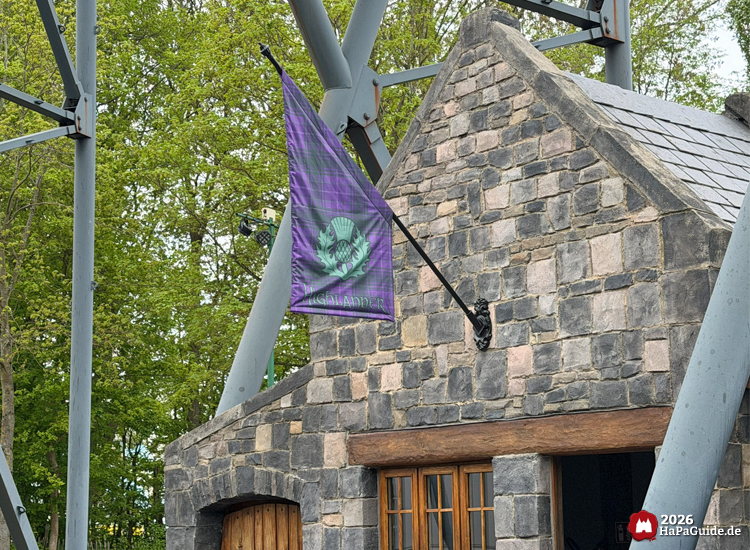 Steingebäude mit Holzfenstern am Highlander im Hansa-Park, an dem eine violett-karierte Flagge mit grünem Distel-Emblem und der Aufschrift Highlander weht