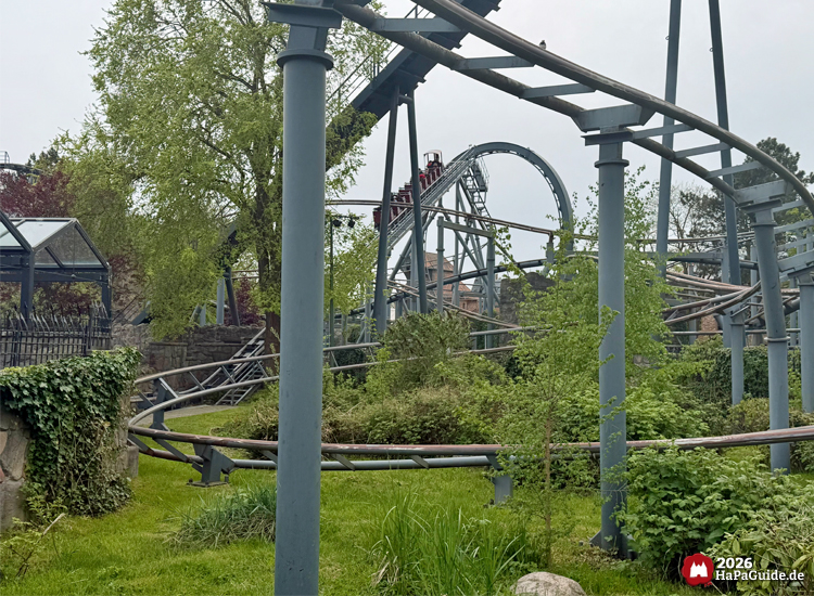 Verschlungene Schienen der Familienachterbahn Royal Scotsman im Hansa-Park, umgeben von dichtem Grün und Bäumen, mit hellgrauen Stahlstützen und einem Looping im Hintergrund an einem leicht bewölkten Frühlingstag