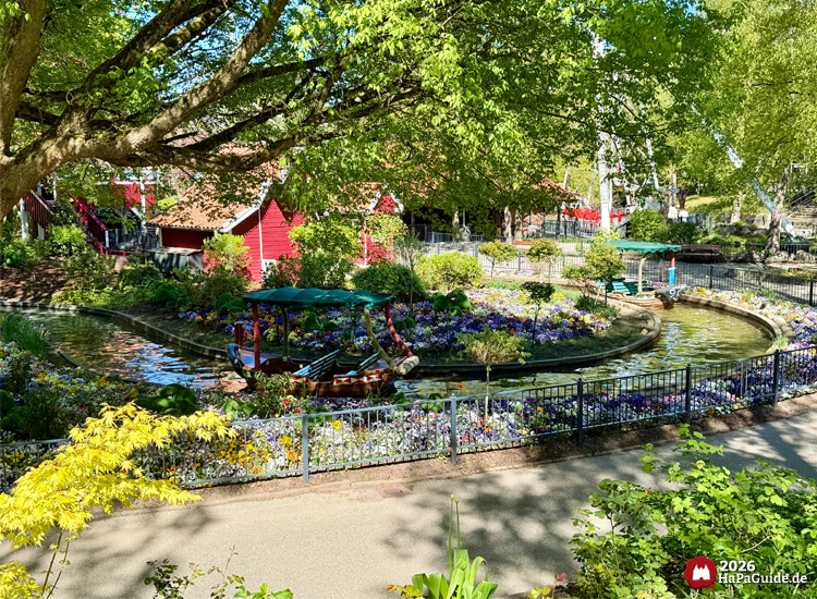 Übersicht über die Blumenmeerbootsfahrt des Hansa-Parks mit dekorativem Boot auf dem Wasserkanal, umgeben von bunten Blumen und roten Gebäuden unter grünem Blätterdach