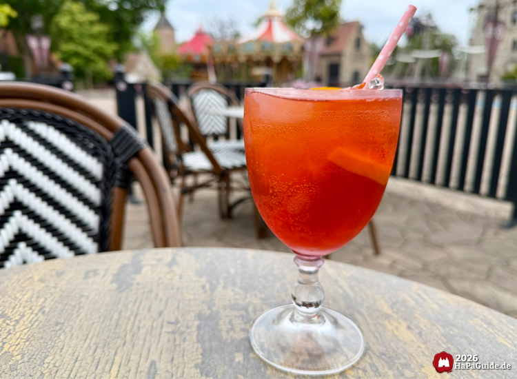Erfrischender Aperol Spritz in einem Glas mit Eiswürfeln und Orangenscheibe auf einem Tisch der Außenterrasse des Café Rosa im Hansa-Park, im Hintergrund Stühle mit schwarz-weißem Muster und das Hanse-Karussell