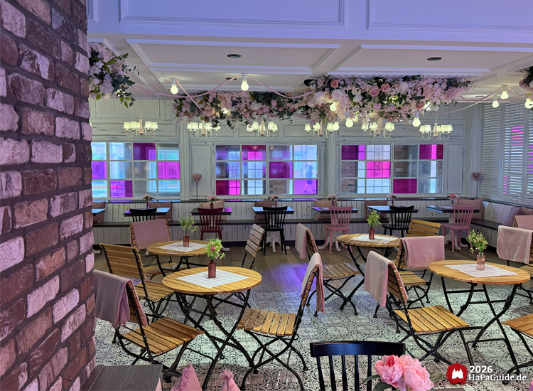 Innenraum des Café Rosa im Hansa-Park mit Holzstühlen, runden Tischen und floraler Deckendekoration aus rosa Blüten, beleuchtet durch warme Lichter und farbige Fenster