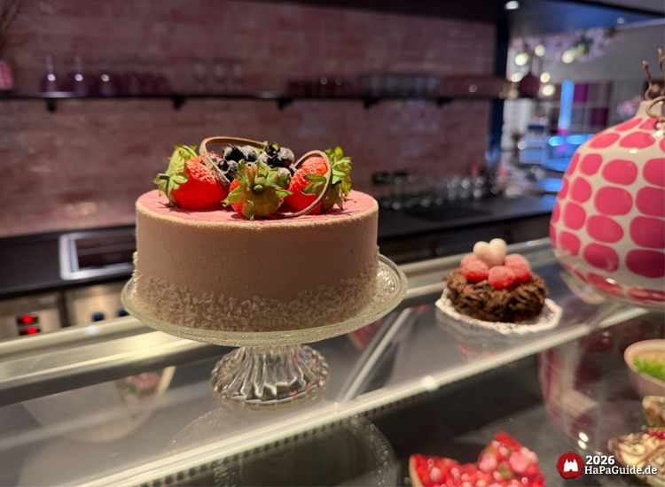 Vitrine im Café Rosa des Hansa-Parks mit rosa Plastiktorte auf Glasständer, dekoriert mit Erdbeeren, Blaubeeren und Schokoladenelementen, daneben weitere kleine Schau-Desserts