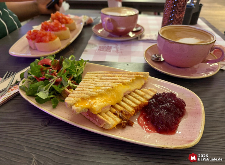 Tisch im Hansa-Park Café Rosa mit Toast-Käse-Schinken-Sandwich, Preiselbeermarmelade, Salat und zwei Tassen Kaffee, im Hintergrund Bruschetta mit Tomaten auf rosa Tellern