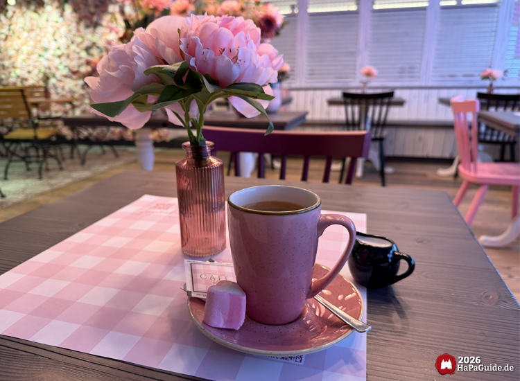 Tasse Americano in rosa Geschirr mit Schaumherz und Vase mit rosa Pfingstrosen auf einem Tisch im Café Rosa des Hansa-Parks, im Hintergrund gemütliche Sitzplätze mit floraler Dekoration
