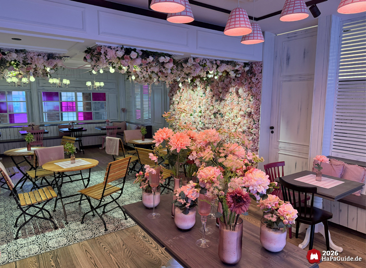 Innenraum des Café Rosa im Hansa-Park mit rosa Blumendekoration, hölzernen Tischen und Stühlen, hellen Wänden und warmem Licht, schafft eine romantische und einladende Atmosphäre