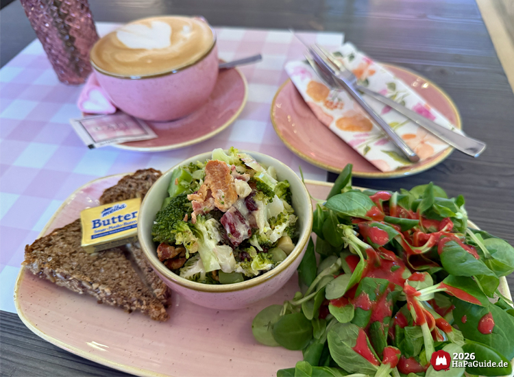 Frischer Brokkolisalat mit Dressing, Vollkornbrot und Butter auf rosa Teller im Café Rosa des Hansa-Parks, daneben eine Tasse Cappuccino mit Herzmuster und rosa Servietten