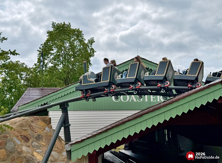 Achterbahn Cornwall Coaster im Hansa-Park fährt über das grün-weiße Stationsgebäude mit roten Dachkanten, während die Züge mit goldenen Zierleisten über die Schienen gleiten, umgeben von Bäumen unter bewölktem Himmel