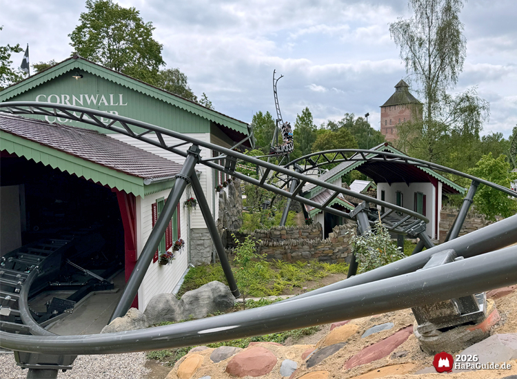 Schwarze Stahlkonstruktion der Achterbahn Cornwall Coaster im Hansa-Park schlängelt sich durch eine thematisierte Landschaft mit weißen Gebäuden, roten Fensterläden und Felsen, im Hintergrund der Saviour-Tower und grüne Bäume