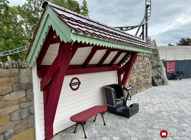 Thematisierte Bushaltestelle am Zugang des Hansa-Park Cornwall Coasters mit rotem Dach, grünem Zierabschluss und weißer Wand mit 'Bus Stop'-Schild, davor eine rote Bank und ein Test-Sitz für die Achterbahn