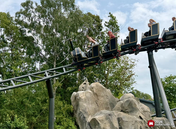 Achterbahn Cornwall Coaster im Hansa-Park fährt über graue Felsen und grüne Bäume, die Wagen im Stil einer schwarzen Dampflokomotive mit goldenen Zierleisten unter blau-grauem Himmel