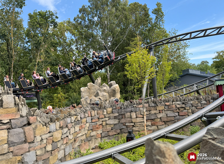 Zug der Cornwall Coaster Achterbahn im Hansa-Park fährt über eine hohe Steinmauer mit Zacken, umgeben von Felsen, Bäumen und grüner Vegetation unter blauem Himmel