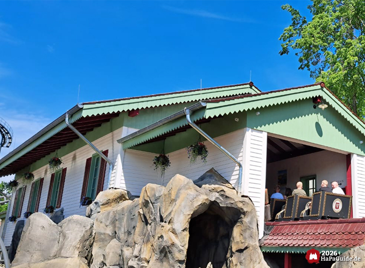 Detailreich gestaltetes Bahnhofsgebäude im Hansa-Park mit weiß-grüner Fassade, Felskulisse und sichtbarem Zug des Cornwall Coasters, in dem Besucher in den hinteren Wagen sitzen