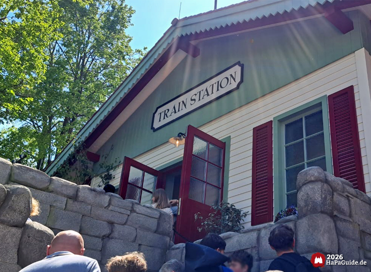 Eingangsbereich des Bahnhofsgebäudes vom Cornwall Coaster mit der Aufschrift „Train Station“ im Hansa-Park, hell gestrichene Fassade mit grünen Zierleisten und roten Fensterläden, davor Besuchende in der Warteschlange bei sonnigem Wetter