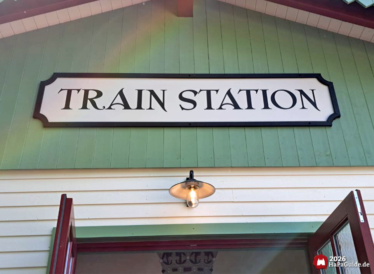 Nahaufnahme des Cornwall Coaster Bahnhofs mit der Aufschrift „Train Station“ im Hansa-Park, hellgrüne und cremefarbene Holzfassade mit rotem Rahmen, beleuchtetem Lampenschirm über dem Eingang und geöffneten Türen