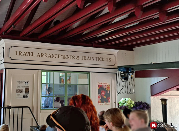 Innenbereich der Cornwall Coaster Station im Hansa-Park mit der Aufschrift „Travel Arrangements & Train Tickets“, grün gerahmtem Schalterfenster, roten Deckenbalken und wartenden Parkgästen vor dem nostalgisch gestalteten Ticketschalter