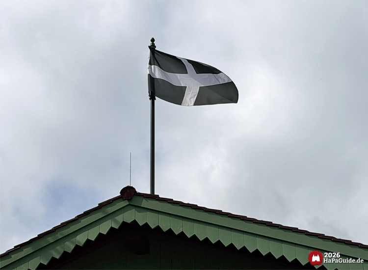 Schwarze Cornwall-Flagge mit weißem Kreuz auf einem grünen Holzgiebel im Hansa-Park vor grauem Wolkenhimmel