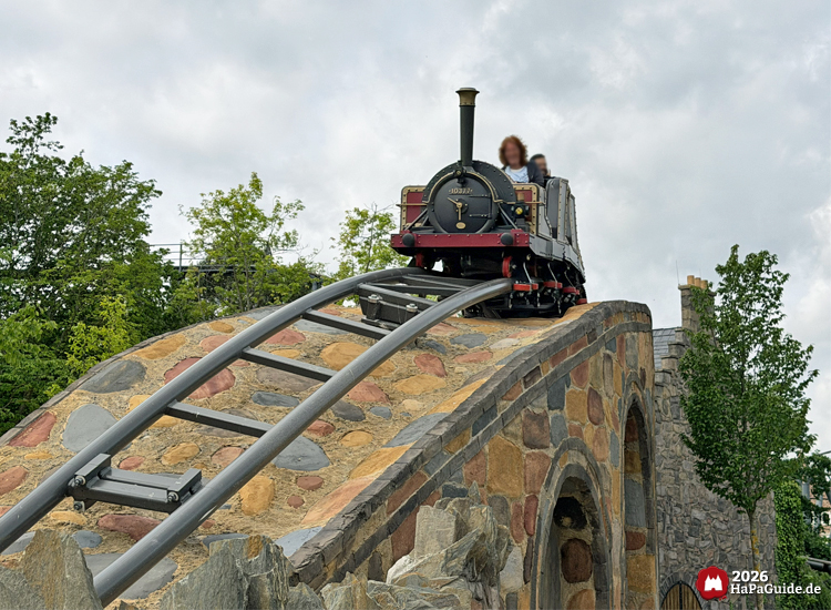 Kleine schwarze Dampflokomotive des Cornwall Coasters mit goldenen Details fährt über eine steinerne Brücke im Hansa-Park, umgeben von grünen Bäumen und grauem Himmel