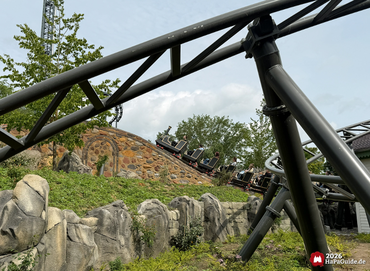 Cornwall Coaster im Hansa-Park mit schwarzen Stahlschienen im Vordergrund und Zug auf einer steinernen Brücke, umgeben von Felsen, Grünpflanzen und blauem Himmel mit Wolken