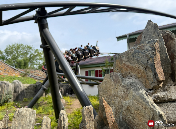 Cornwall Coaster im Hansa-Park fährt hinter dunklen Stahlschienen und an Felsen vorbei, mit Fahrgästen in schwarzen Wagons vor einem Gebäude mit grünem Dach und roten Fensterläden unter blauem Himmel