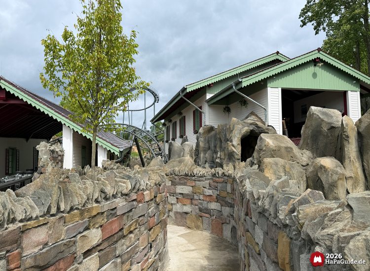 Steinweg mit Felsen und gemauerter Wand führt durch thematisierte Landschaft des Cornwall Coasters im Hansa-Park, flankiert von weißen Gebäuden mit grünen Dächern und roten Fensterläden, im Hintergrund Achterbahnschienen und ein Baum unter grauem Himmel