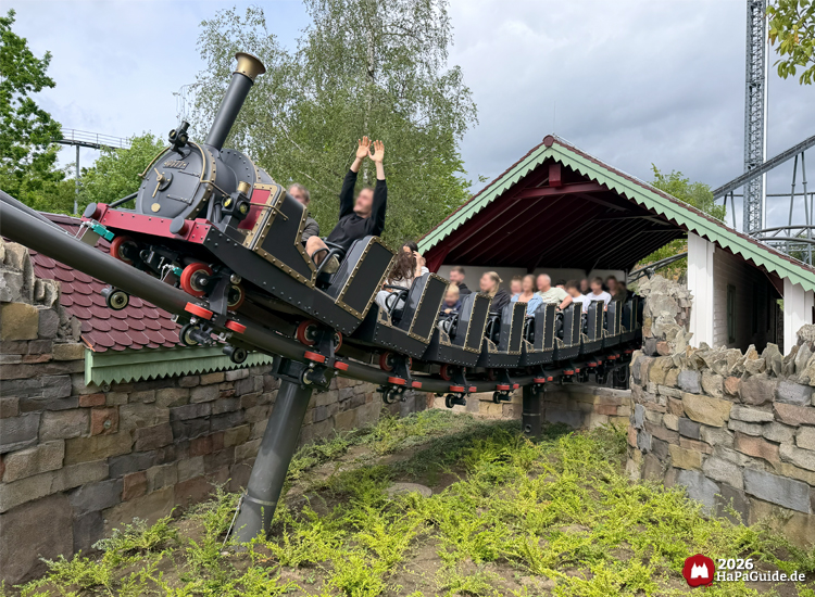 Schwarze Dampflokomotive als Achterbahnzug Cornwall Coaster im Hansa-Park fährt aus einem überdachten Tunnel mit grüner Dachkante und roten Balken, umgeben von Steinmauern und grünen Sträuchern, während Fahrgäste die Arme in die Luft strecken