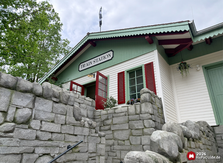 Helle Bahnhofsfassade des Cornwall Coasters im Hansa-Park mit grünem Dachrand, roten Fensterläden und offenem Eingang unter dem Schild 'Train Station', umgeben von grauen Steinmauern und grünen Bäumen unter leicht bewölktem Himmel
