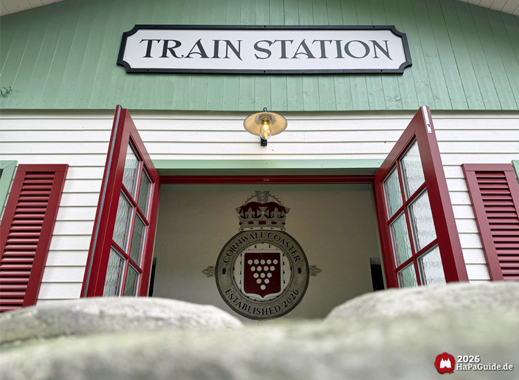Eingang des Cornwall Coaster Bahnhofsgebäudes im Hansa-Park mit geöffneten roten Türen unter dem Schild 'Train Station', darüber eine Lampe und im Inneren ein Wappen mit der Aufschrift 'Cornwall Coaster Established 2016', eingerahmt von grünen und roten Fassadendetails