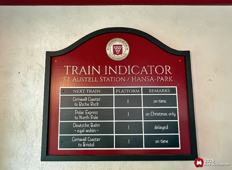 Dekorative Anzeigetafel im Bahnhof des Hansa-Park Cornwall Coasters mit der Überschrift 'Train Indicator – St. Austell Station / Hansa-Park', rotem Hintergrund und Einträgen zu Zügen wie 'Cornwall Coaster', 'Polar Express' und 'Deutsche Bahn', jeweils mit Plattform- und Zeitangaben