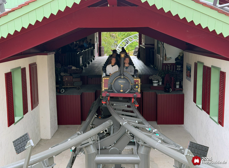 Cornwall Coaster im Hansa-Park mit schwarzer Lokomotive fährt aus dem überdachten Bahnhof mit rotem Dach und grünen Zierleisten, während Fahrgäste in den Wagen sitzen und die schwarze Schienenkonstruktion sich verzweigt
