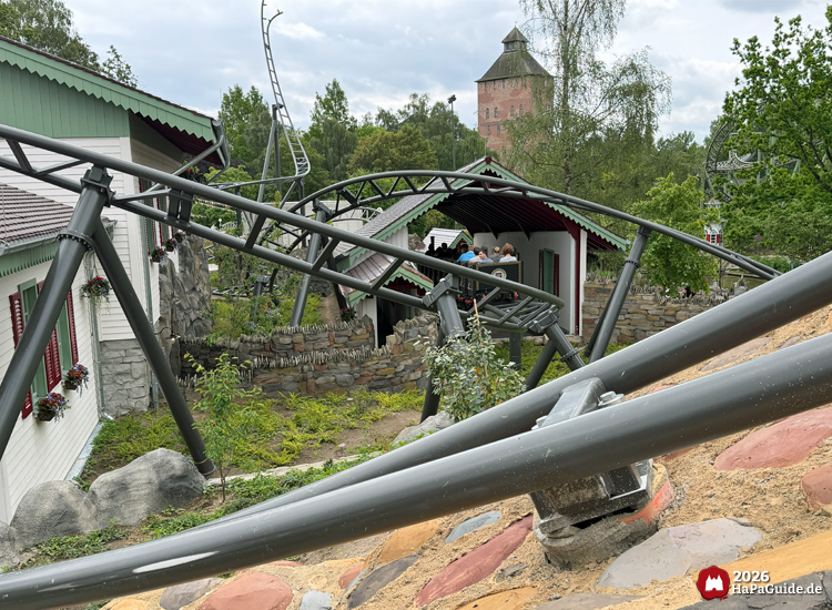 Der Cornwall Coaster im Hansa-Park mit schwarzer Stahlkonstruktion schlängelt sich zwischen weißen Gebäuden mit grünen Dächern und roten Fensterläden, umgeben von Felsen und Bäumen, im Hintergrund der Saviour-Tower unter grauem Himmel