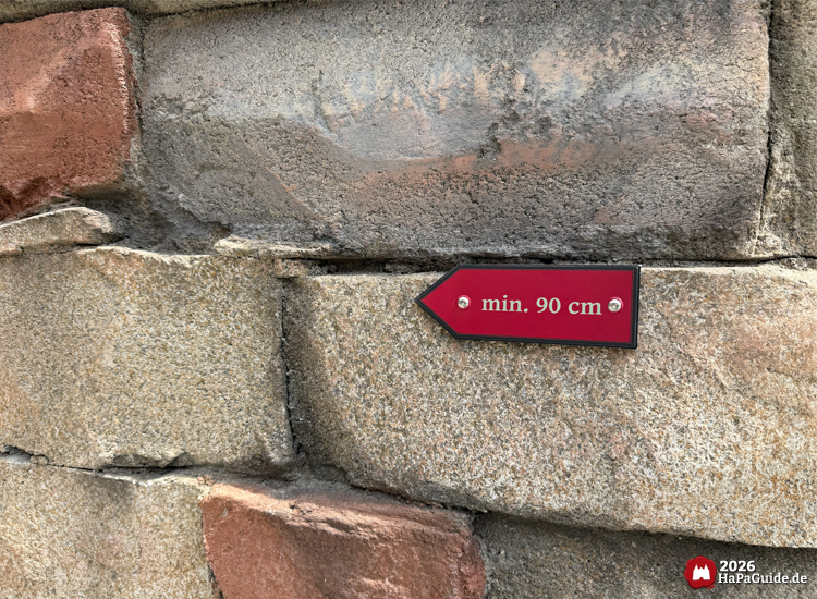 Rotes Schild mit weißer Aufschrift 'min. 90 cm' an einer gemauerten Steinwand des Hansa-Park Cornwall Coasters, Hinweis auf Mindestgröße für die Achterbahn