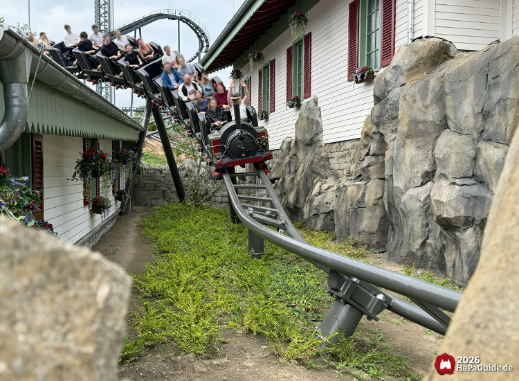 Die schwarze Dampflokomotive des Cornwall Coasters im Hansa-Park fährt zwischen weißen Gebäuden mit roten Fensterläden und Felsen hindurch, umgeben von grüner Vegetation und grauer Stahlstrecke