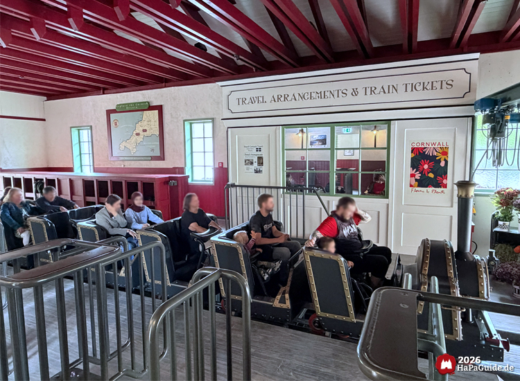 Innenbereich der Cornwall Coaster Station im Hansa-Park mit roten Deckenbalken, Fahrgästen im Zug und Schildern wie 'Travel Arrangements & Train Tickets' sowie einer Cornwall-Karte an der Wand