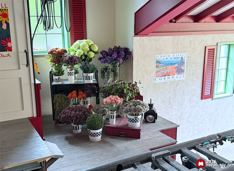 Dekorative Blumenarrangements in weißen Töpfen mit schwarz-weißem Muster im Bahnhof des Hansa-Park Cornwall Coaster, daneben ein Poster mit der Aufschrift 'Flower Market Cornwall' und rote Fensterläden