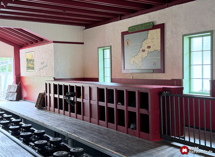 Thematisierte Cornwall Coaster Station im Hansa-Park mit rotem Holzdekor, einer Karte der 'Cornish Riviera' zwischen grünen Fenstern und einem Reiseplakat 'Visit Lanhydrock' an der Wand