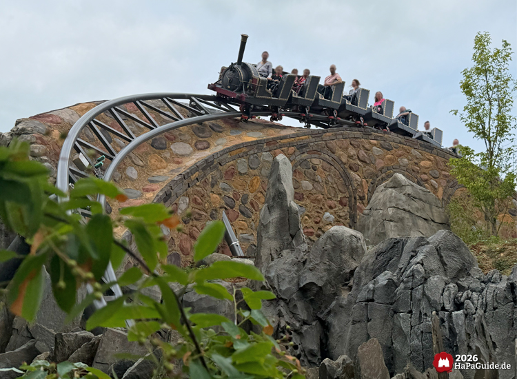 Cornwall Coaster im Hansa-Park mit schwarzer Lokomotive fährt über eine steinerne Brücke aus bunten Felsen, umgeben von grauen Felsformationen und grüner Vegetation unter bewölktem Himmel