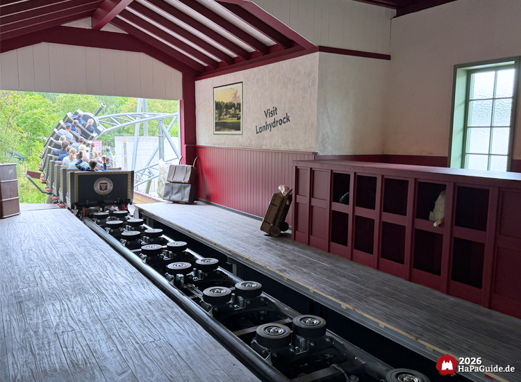 Der Cornwall Coaster im Hansa-Park fährt aus der Station mit rotem Dach und Wandaufschrift 'Visit Lanhydrock', im Vordergrund Reibräder und rote Gepäckfächer