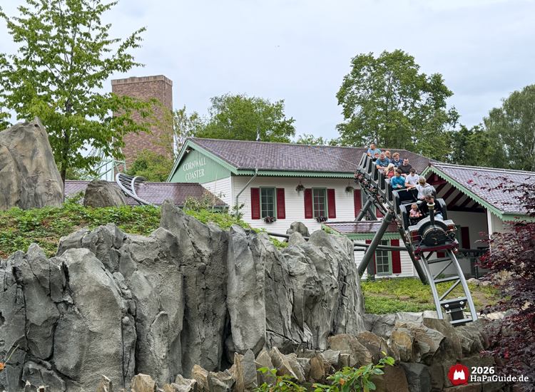 Achterbahn Cornwall Coaster im Hansa-Park fährt aus einem weißen Gebäude mit roten Fensterläden und grünem Dach, umgeben von Felsen und Bäumen unter bewölktem Himmel