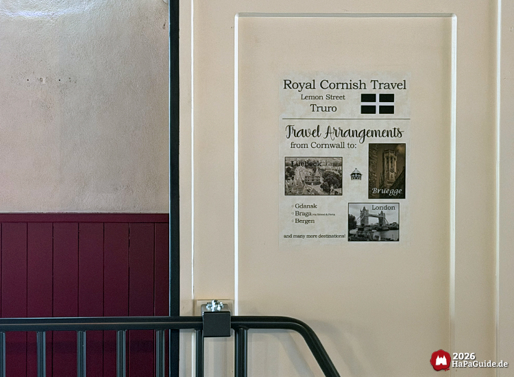 Reiseposter im Bahnhof des Hansa-Park Cornwall Coasters mit der Aufschrift 'Royal Cornish Travel – Lemon Street Truro', zeigt Reiseziele wie Lübeck, Brügge und London auf einer cremefarbenen Wand mit rotem Sockel