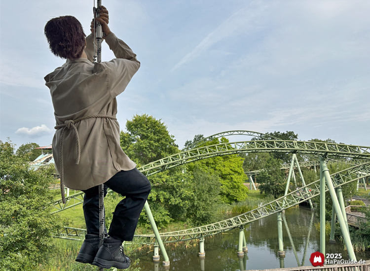 Figur eines Minenarbeiters, der an einem Seil am Minenförderturm der Crazy Mine hängt, mit Blick auf die grünen Schienen zum Schwur des Kärnan im Hansa-Park, umgeben von Bäumen und blauem Himmel