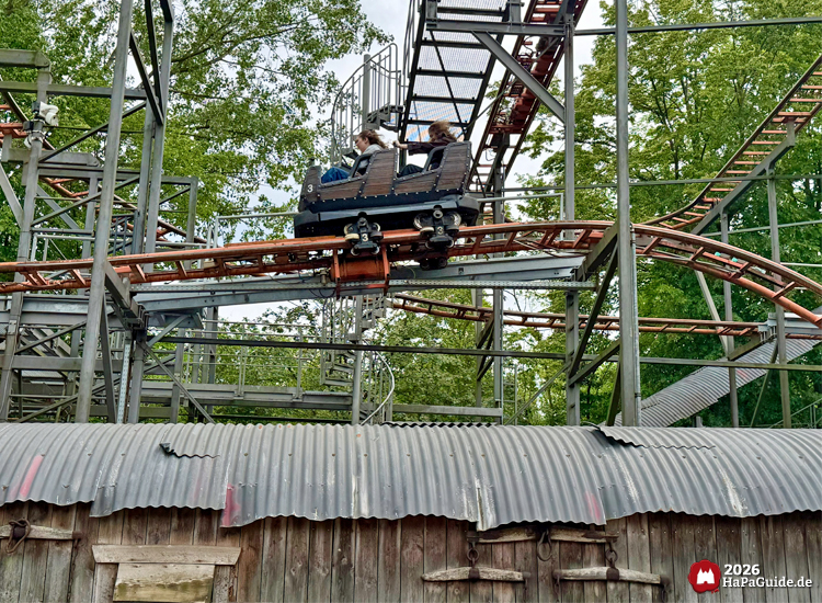 Achterbahn Crazy Mine im Hansa-Park mit roten Schienen und grauer Stahlkonstruktion, Wagen mit Fahrgästen fährt in eine Bremse, im Vordergrund ein Wellblechdach