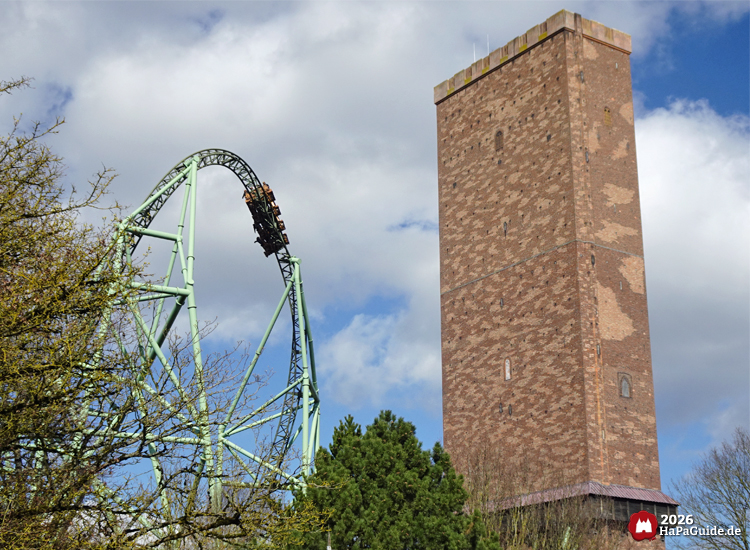 Achterbahn Der Schwur des Kärnan im Hansa-Park mit grünem Herzelement und hohem Backsteinturm, Zug befindet sich auf der Spitze der Strecke unter blauem Himmel mit Wolken
